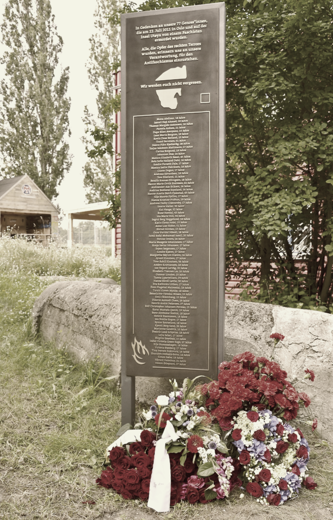 Das Denkmal für die Ermordeten am 22. Juli 2011 in Oslo und Utøya steht in der Mitte des Bildes und im Hintergrund sieht man ein Gebäude mit einem Falken-Logo drauf.